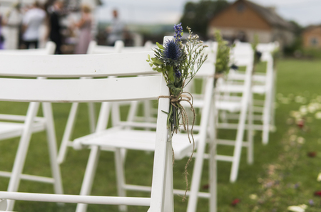 White wedding chairs decorated with fresh flowers on a green grass. Empty wooden chairs for guests on green lawn in the garden prepared for wedding ceremony.の写真素材