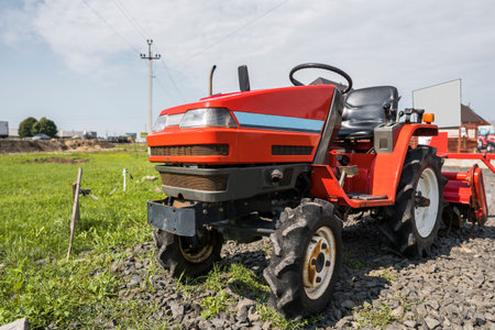 A small mini red tractor stands on a farm yard on green grass and waits for work to begin.