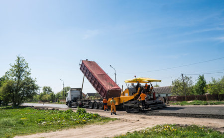 Worker operating asphalt paver machine during road construction and repairing works. A paver...