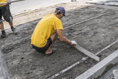 Builder in his orange gloved and yellow shirt prepares the surface before laying the paving stones with the ruler of the level. Sidewalk building.の写真素材