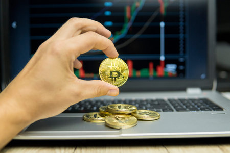 Male businessman hand holding bitcoin on a background of growth graph on a screen of laptop and pile...