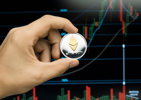 Male businessman hand holding Ethereum coin on a background of growth graph on a screen of laptop....