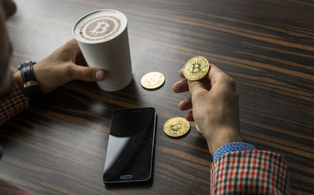 Rich man holding a bitcoin coin and drinking a coffee with a phone and gold coins of bitcoin on a table. Profit from mining of bitcoins.の写真素材