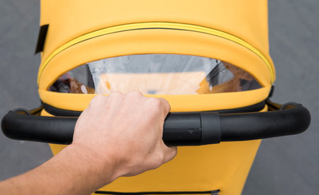 Young woman strolling a carriage outdoor. Closeup shot of male hands with yellow stroller handle....