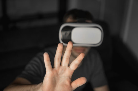 Young bearded man wearing headset of virtual reality in office sitting on a sofa and trying to touch...