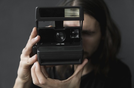 Handsome young bearded man with a long hair and in a black shirt holding vintage old-fashioned film camera on a black background and looking in camera viewfinder.の写真素材