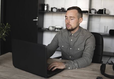 Young businessman or student in a shirt sitting against monitor of computer. Working on a pc at a table in the office with a thoughtful expression. Young businessman working on his laptop in office.の写真素材