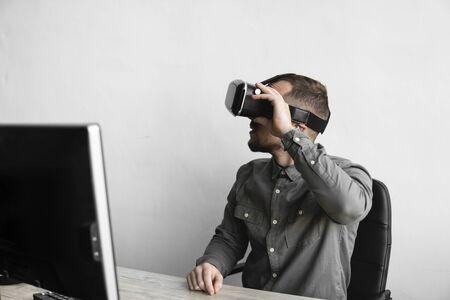 Young bearded man sitting against computer with virtual reality glasses. Modern technologies. The concept of future technology.の写真素材