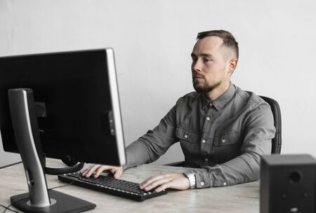 Young businessman or student in a shirt sitting against monitor of computer. Working on a pc at a table in the office with a thoughtful expression. Young businessman working on his laptop in office.の写真素材