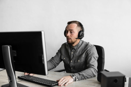 Young businessman or student in a shirt sitting against monitor of computer in headphones. Working on a pc at a table in the office with a thoughtful expression.の写真素材