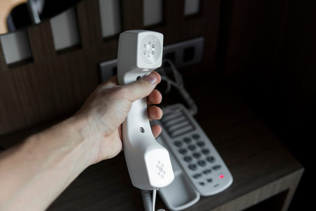 Mans hand holding handset of white vintage telephone with grey battoms on a brown table in hotel.