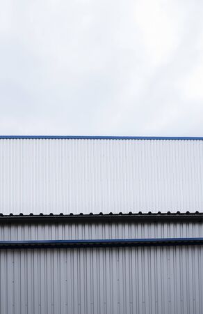 Metal corrugated sheets on a building with a blue metal corners. White aluminium metal corrugated roof or wall sheets against cloudy sky background on a factories and industrial buildings.の写真素材
