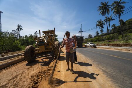 THAILAND, PHUKET - December 12, 2018: Women working on a road construction site. Grader is working on road construction. Grader industrial machine on construction of new roads. Heavy duty machinery working on highway.のeditorial素材