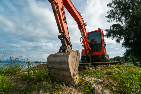 Small orange excavator on a ground against blue sky and sea for a works on construction site. Small...