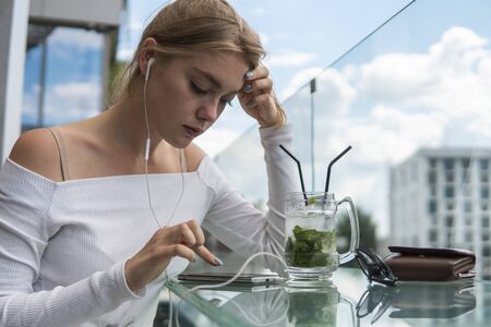 Cheerful blond woman blogger with cocktail is reading incoming sms message on smartphone connected to free wifi in cafe. Positive young student watching broadcasting online on modern mobile phone.の写真素材