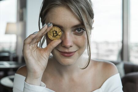 Close up fun emotional young female is holding bitcoin in front eye. Online virtual future currency concept. Beauty woman head with Bitcoin money in eye. Digital cryptocurrency bitcoin.の写真素材