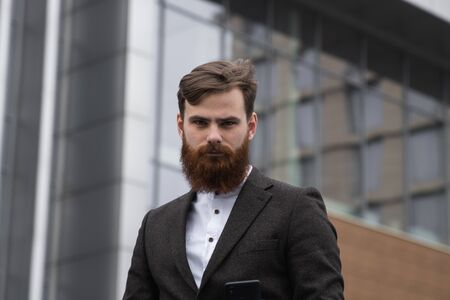 Portrait of an handsome businessman wearing jacket over office building in financial district outdoors. Young bearded Businessman outdoor. Successful entrepreneur dressed in formal wear.の写真素材