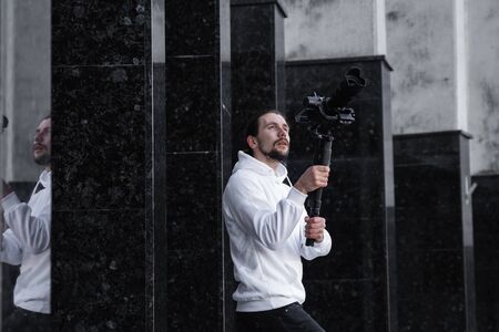 Young Professional videographer holding professional camera on 3-axis gimbal stabilizer. Pro equipment helps to make high quality video without shaking. Cameraman wearing white hoodie making a videos.の写真素材