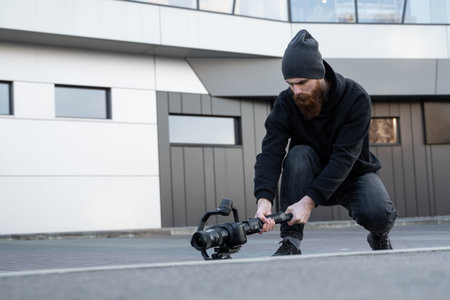Bearded Professional videographer in black hoodie holding professional camera on 3-axis gimbal...