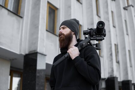 Bearded Professional videographer in black hoodie holding professional camera on 3-axis gimbal stabilizer. Filmmaker making a great video with a professional cinema camera. Cinematographer.の写真素材