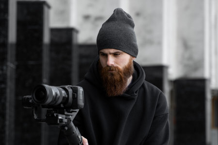 Bearded Professional videographer in black hoodie holding professional camera on 3-axis gimbal...