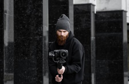 Bearded Professional videographer in black hoodie holding professional camera on 3-axis gimbal stabilizer. Filmmaker making a great video with a professional cinema camera. Cinematographer.の写真素材