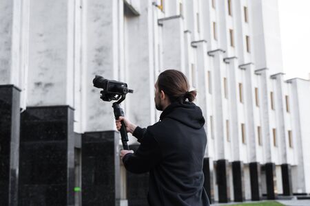 Young Professional videographer holding professional camera on 3-axis gimbal stabilizer. Pro equipment helps to make high quality video without shaking. Cameraman wearing black hoodie making a videos.の写真素材
