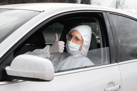 Man in protective suit, medical mask and rubber gloves for protect from bacteria and virus is sitting in a car and showing a thumb up. Quarantine, world pandemic, covid 19, coronavirus, infection.の写真素材