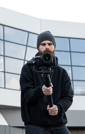 Bearded Professional videographer in black hoodie holding professional camera on 3-axis gimbal...