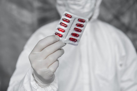 Doctor in white protective suit, medical mask and rubber gloves is holding a red pills as a...