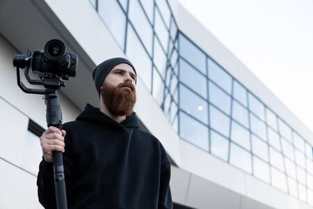 Bearded Professional videographer in black hoodie holding professional camera on 3-axis gimbal stabilizer. Filmmaker making a great video with a professional cinema camera. Cinematographer.の写真素材
