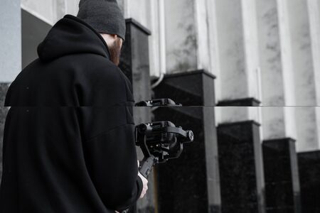 Bearded Professional videographer in black hoodie holding professional camera on 3-axis gimbal...