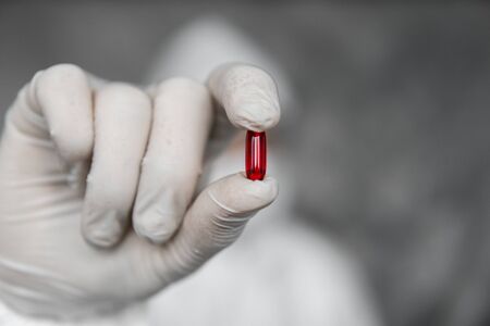 Doctor in white protective suit, medical mask and rubber gloves is holding a red pill as a medications from coronavirus pandemic threat. Epidemic, pandemic of coronavirus covid 19.の写真素材