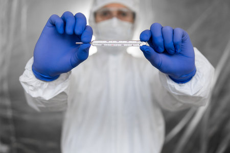Doctor in white protective suit, medical mask and rubber gloves is holding a thermometer in...