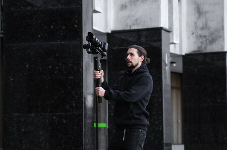 Young Professional videographer holding professional camera on 3-axis gimbal stabilizer. Pro equipment helps to make high quality video without shaking. Cameraman wearing black hoodie making a videos.の写真素材