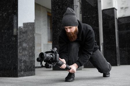 Bearded Professional videographer in black hoodie holding professional camera on 3-axis gimbal stabilizer. Filmmaker making a great video with a professional cinema camera. Cinematographer.の写真素材