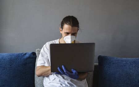 Man in medical mask and rubber gloves sits at home and works with laptop at the sofa during quarantine. Designer, artist, architect, businessman at remote work in a pandemic covid. Coronavirus.の写真素材