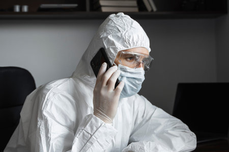 Man in protective suit, medical mask and rubber gloves sits at home and speaking on a phone with...