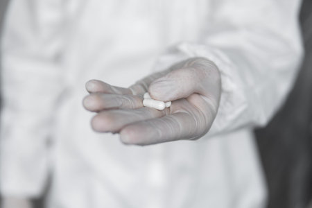 Doctor in white protective suit, medical mask and rubber gloves is holding a white pills as a...
