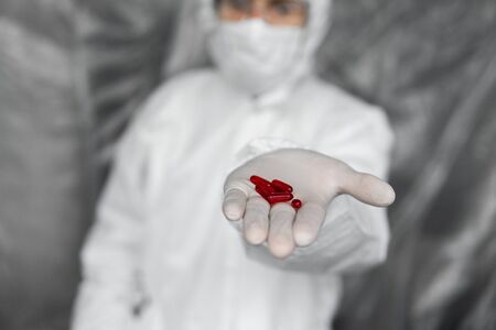 Doctor in white protective suit, medical mask and rubber gloves is holding a red pills as a medications from coronavirus pandemic threat. Epidemic, pandemic of coronavirus covid 19.の写真素材