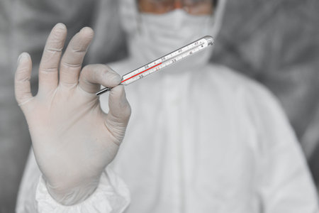 Doctor in white protective suit, medical mask and rubber gloves is holding a thermometer in...