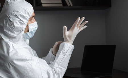Man in protective suit and medical mask is wears rubber gloves at his working place at home during...
