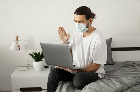 Man in medical mask is working from home with a laptop and speaking to his friends or business partners via webcam because of coronavirus.の写真素材