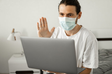 Man in medical mask is working from home with a laptop and speaking to his friends or business...