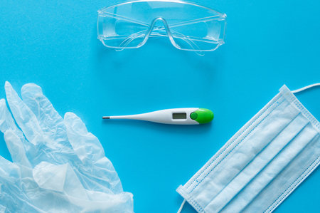 Medical protective mask, rubber gloves, thermometer and protective glasses lie on a blue background....