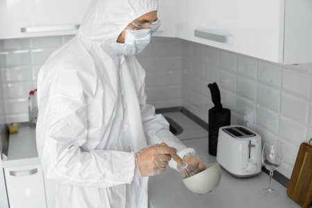 Man in white protective suit and in face medical mask cooks in the white kitchen at home during...