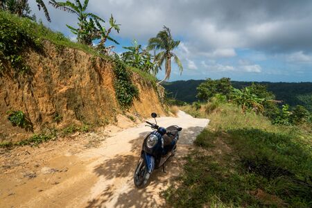 Blue scooter on the dirt road on a hill in tropical jungle with a scenic view on a mountains in a sunny day. Adventure and traveling concept.の写真素材