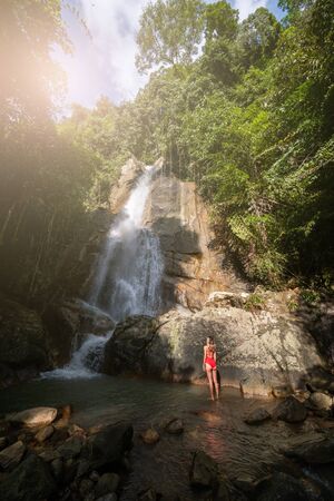 Beautiful young slim blonde woman with curly hair, wearing in red swimsuit is enjoy in lagoon of huge tropical waterfall in jungle. Travel concept. Sexy woman wearing red swimsuit in the jungle.の写真素材