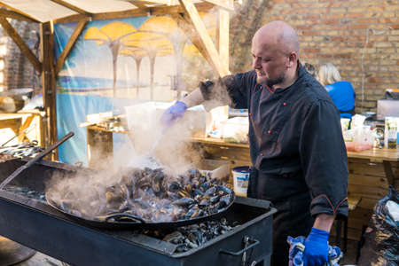 UKRAINE, LUTSK - June 5, 2019: Man is cooking a fresh mussels in shells in large metallic grill pan on a food fest.のeditorial素材