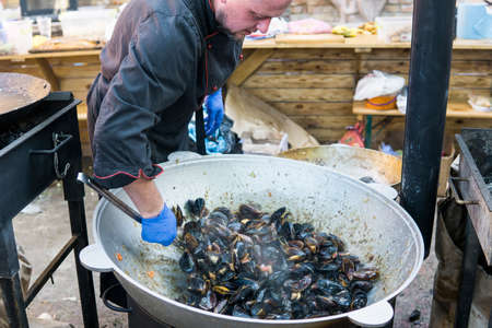 UKRAINE, LUTSK - June 5, 2019: Man is cooking a fresh mussels in shells in large metallic grill pan on a food festのeditorial素材